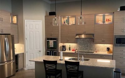 A modern kitchen remodel with wooden cabinets, stainless steel appliances, and recessed lighting. A granite island with a sink and two high chairs is centrally located. The cabinets have glass displays with lighting, and pendant lights hang above the island.