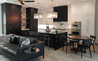 A modern kitchen and living area with black and white cabinetry, a large island with bar stools and pendant lights, a round dining table, and a black leather sofa with accent pillows. The remodel in Crystal River, FL features stainless steel appliances and a light wood floor.