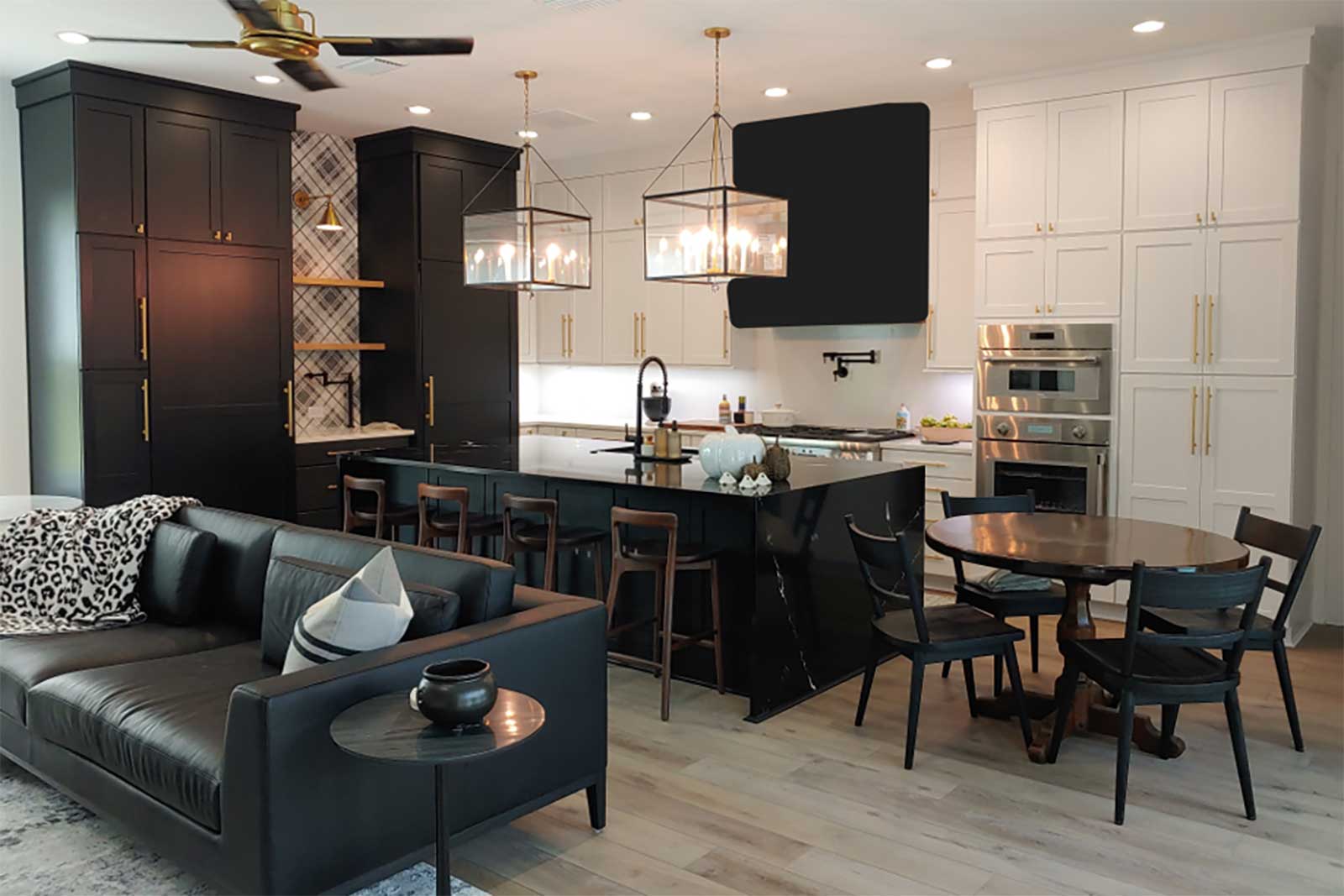 A modern kitchen and living area with black and white cabinetry, a large island with bar stools and pendant lights, a round dining table, and a black leather sofa with accent pillows. The remodel in Crystal River, FL features stainless steel appliances and a light wood floor.