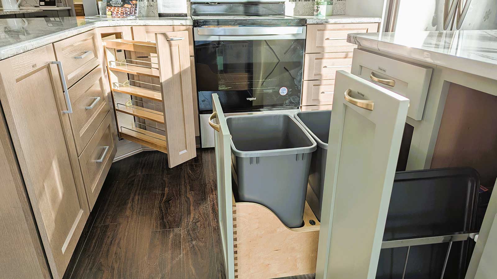 A modern kitchen with light wood cabinets. Open cabinets reveal a pull-out spice rack and a pull-out double trash bin. A black oven is central, and the floor is dark wood. Countertops have various kitchen items. Natural light brightens the space.