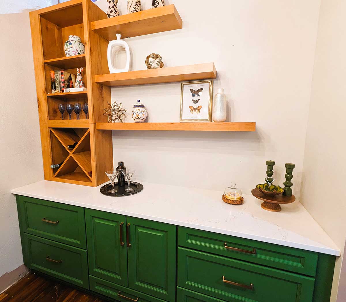 Green cabinets with sleek, white countertops feature wooden shelves above displaying ceramics, a butterfly frame, candles, and wine glasses. A wooden wine rack seamlessly integrates into the shelving, while the cabinet holds a cocktail shaker and glasses on a round tray.