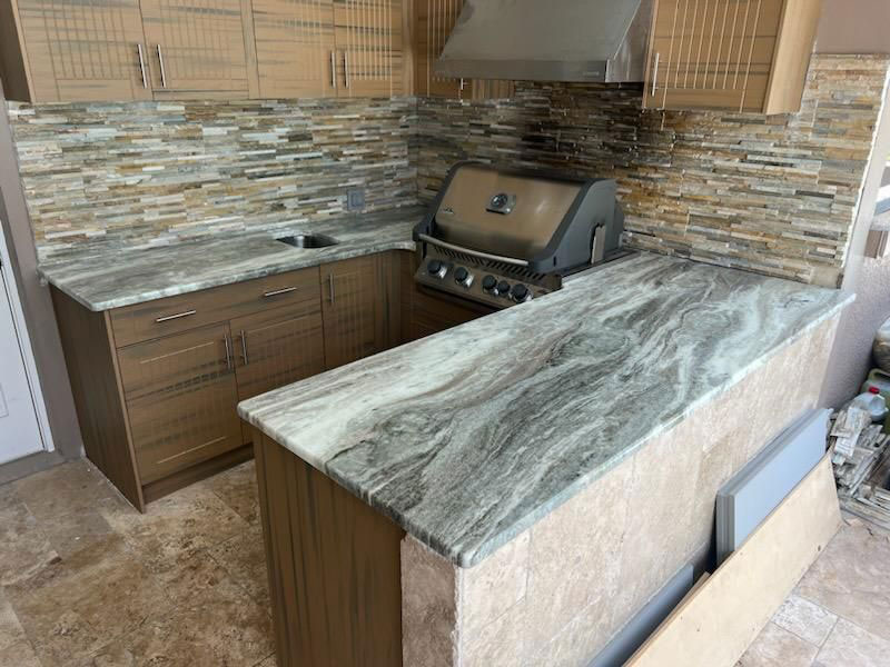 Wolfman-Family-Outdoor-Kitchen Modern outdoor kitchen remodel featuring brown cabinets, marble-patterned countertops, a built-in grill, small sink, and tiled stone backsplash. The scene highlights a clean, newly installed setup.