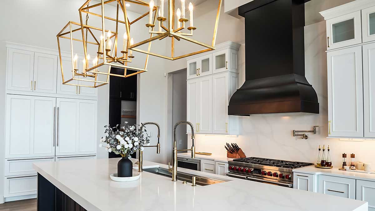 Modern kitchen with white cabinets, a large marble island, gold geometric light fixtures, double sinks with tall faucets, elegant kitchen stove hoods in black, and stainless steel appliances. A vase with flowers sits on the island.
